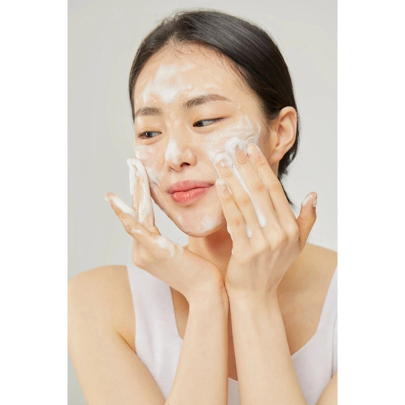 Jeune femme asiatique se lavant le visage avec un gel crémeux blanc, mousse généreuse sur les joues, sourire léger.